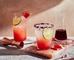 Two glasses of hibiscus paloma with a pitcher of simple syrup, shown with a jigger and skewers.