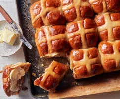 Hot cross buns on a baking sheet with one removed, sliced, and buttered