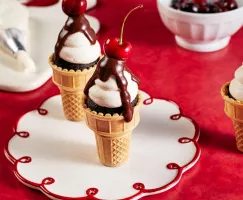 Chocolate cupcakes in classic ice cream cones decorated with icing, chocolate ganache, and topped with a cherry served on a red 