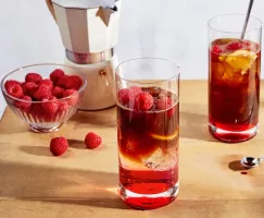 Two glasses of iced raspberry espresso tonic on a wooden table shown with a bowl of raspberries and an espresso maker