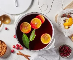 A pot of mulled wine with orange slices and bay leaves shown with sliced ginger, cranberries, and brown sugar