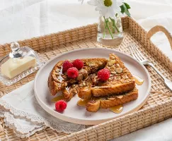 Oat and almond-crusted French toast on a rattan serving tray with butter in a glass dome and flowers in a vase