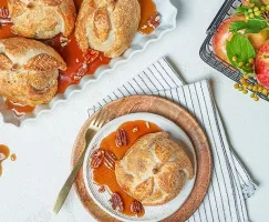 Three apple dumplings on a tray with apple cider syrup, and one on a plate