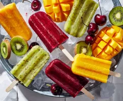 A variety of frozen fruit paletas in a bucket of ice served with kiwis, cherries, and mangoes