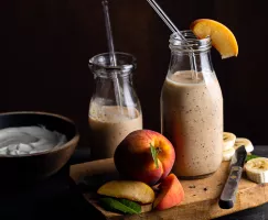 Two single serving bottles of banana peach Earl Grey smoothie with glass straws, with a cutting board and fruit