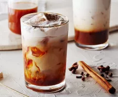 Glass of pumpkin spice cold brew with swirls of cream and cinnamon sprinkles shown with a stick of cinnamon and cloves