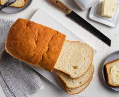 A partially sliced loaf of bread on a cutting board, with a bread knife and one slice buttered on a plate