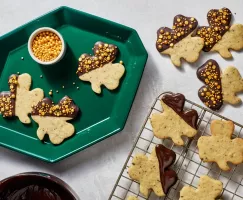 Shamrock-shaped mint cookies dipped in chocolate and decorated with gold sprinkles on a green tray and a wire cooling rack