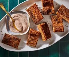 Seven snickerdoodle cookie bars on a white platter with a dish of cinnamon sugar and a gold spoon, shown on a green tile counter