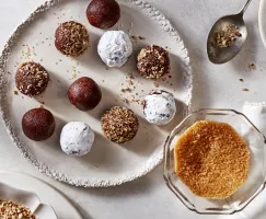 Spiced bourbon balls on a platter, topped with icing sugar, crushed nuts, and turbinado sugar, shown with bowls of toppings.