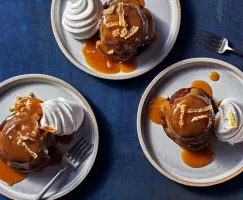 Top down view of three sticky toffee puddings on plates, each with a dollop of whipped cream and toffee sauce.