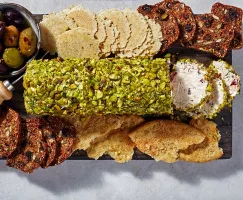 A cream cheese log coated in crushed pistachios on a serving board, surrounded by crisps and crackers and a bowl of olives