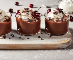 Three vegan chocolate puddings topped with whipped cream, coconut flakes, cacao nibs, and cherries, served on a cutting board