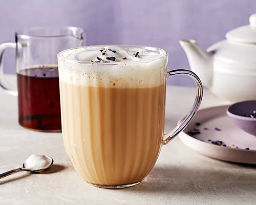 A glass mug of London Fog Latte with milk foam, shown with a pitcher of vanilla syrup, a tea pot, a stirring spoon, and a purple bowl of loose Earl Grey tea on a plate