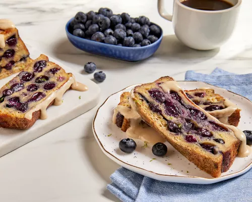 Two slices of loaf pan French toast with blueberries drizzled with maple glaze and shown with a cup of coffee, a dish of blueberries, and the partially sliced loaf 