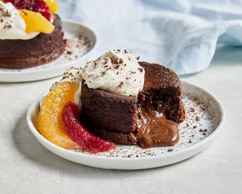 Two personal-size molten chocolate lava cakes on white plates, topped with whipped cream and cocoa powder, garnished with orange and blood orange segments, one shown with a piece missing and chocolate sauce spilling out from the centre.