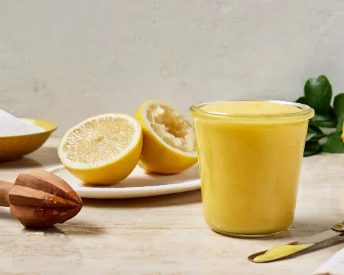 A glass jar full of lemon curd shown with a bisected lemon with one half juiced, a lemon juicer, and a bowl of granulated sugar.