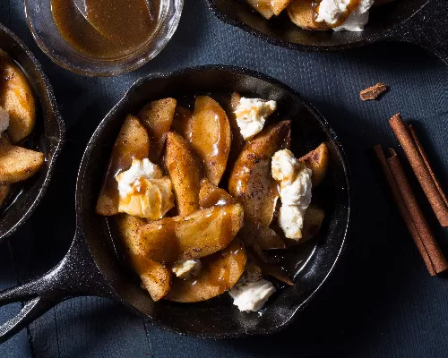 A cast iron skillet of dessert poutine including sliced apples, caramel sauce and sweetened cream