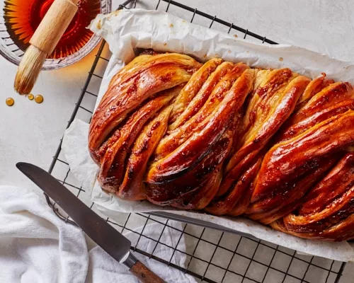 Spiced apple babka in a baking pan on a cooling rack shown with a bowl of honey-brown sugar syrup