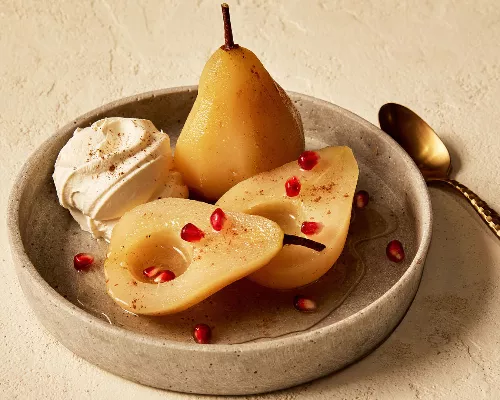 Three halves of white wine poached pears shown in a shallow dish, served with a dollop of whipped cream and sprinkled with cinnamon and pomegranate arils, and shown with a gold spoon.