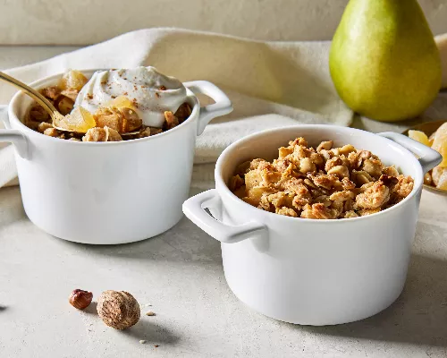 Two white single-serving bowls of pear ginger crumble, one with a dollop of whipped cream and a gold spoon, shown with a whole pear and a small dish of ginger pieces.