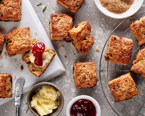 Vanilla rosemary date scones on a platter and a cooling rack shown with dishes of butter and jelly and a bowl of turbinado sugar