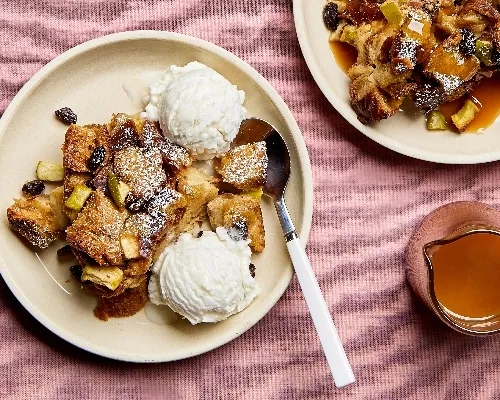 A serving of apple bread pudding on a plate with visible raisins and apple pieces, served with vanilla ice cream, shown with a second plate of bread pudding with butterscotch sauce and a glass pitcher of sauce.