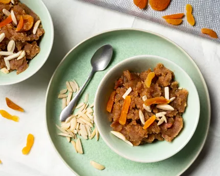 Bowl of whole wheat halwa on a green platter with a spoon and sliced almonds