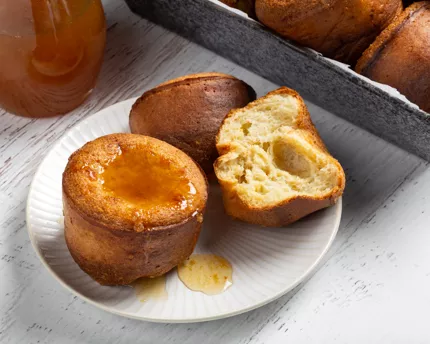 Two lemon spelt popovers on a plate, one cut in half, with lemon syrup on top and a pitcher of lemon syrup behind